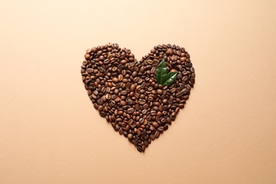 Heart shaped pile of coffee beans and fresh green leaves on light orange background, top view