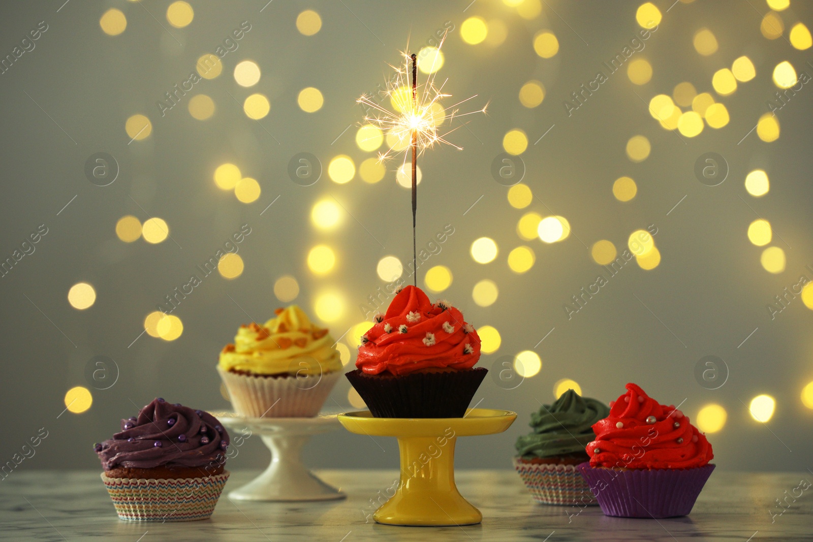 Photo of Different colorful cupcakes and one with sparkler on table against blurred lights