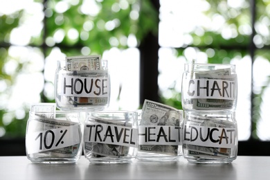 Glass jars with money for different needs on table against blurred background