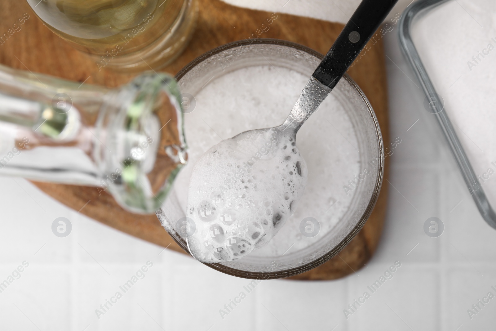 Photo of Pouring vinegar into spoon with baking soda over bowl at white tiled table, top view