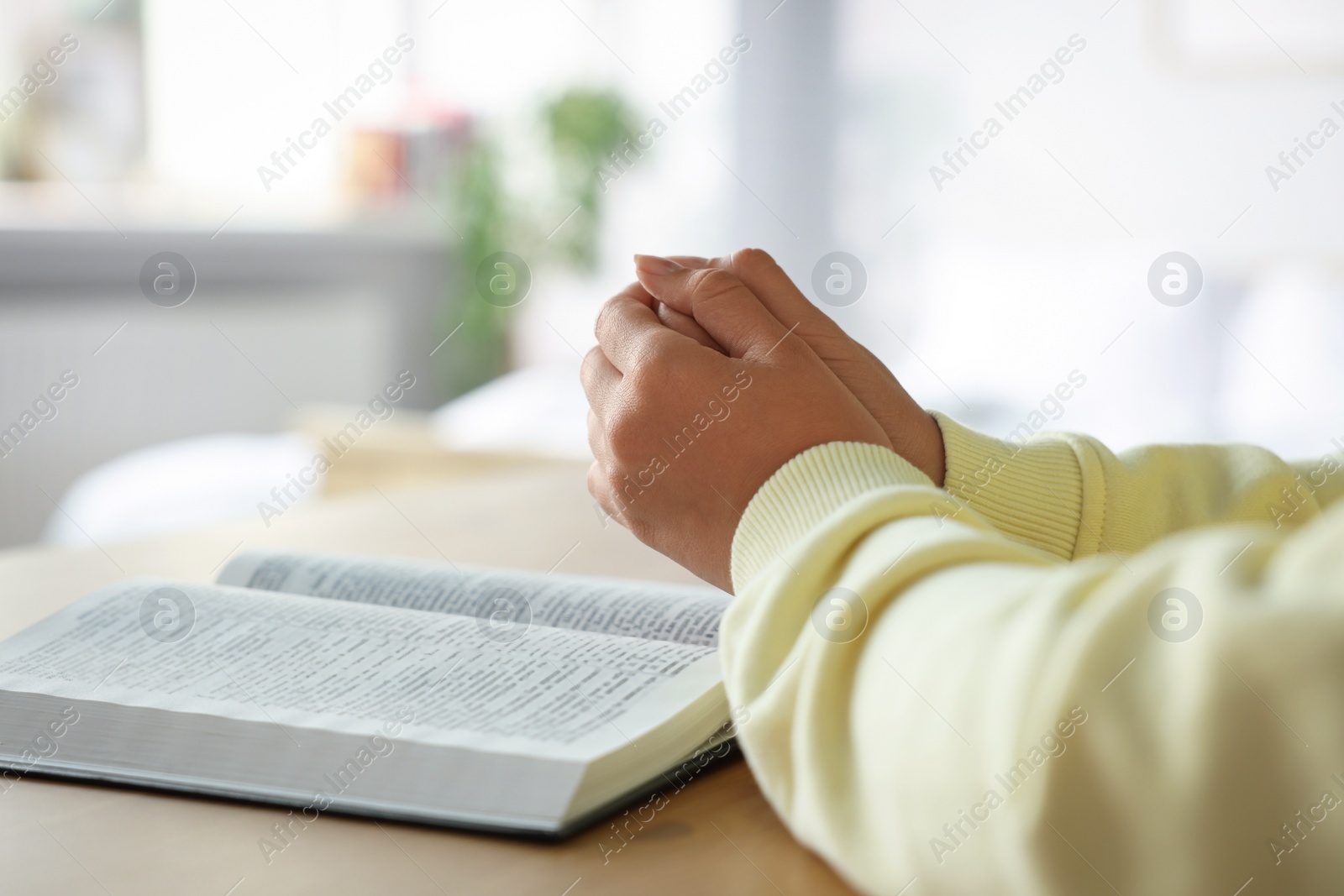 Photo of Woman holding hands clasped while praying at wooden table with Bible indoors, closeup. Space for text