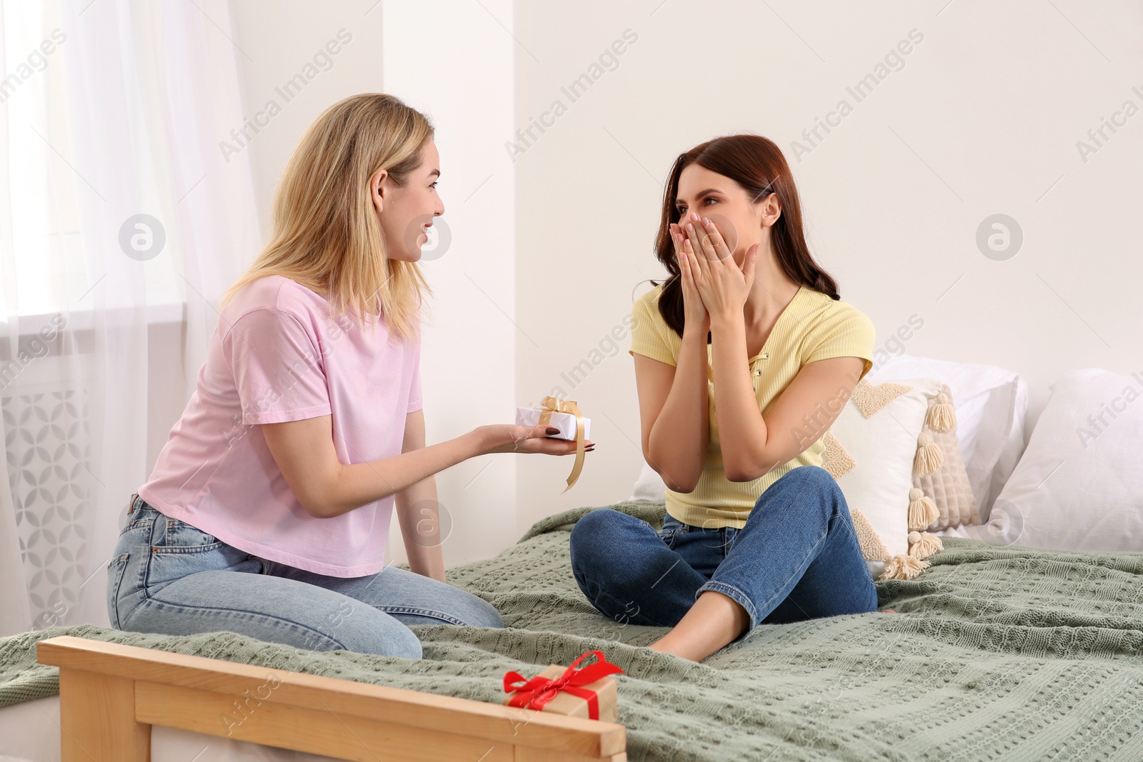 Photo of Smiling young women presenting gifts to each other on bed at home