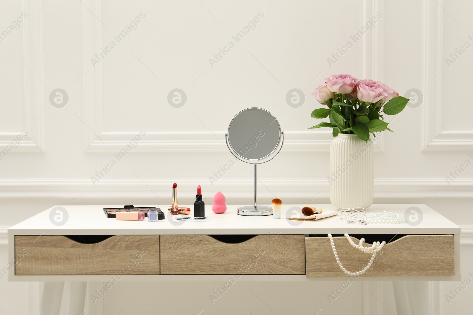 Photo of Mirror, cosmetic products, jewelry and vase with pink roses on white dressing table in makeup room