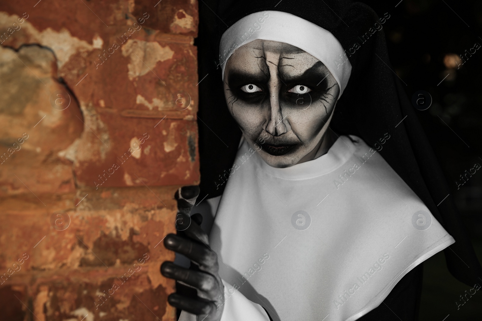 Photo of Scary devilish nun hiding behind red brick wall outdoors. Halloween party look