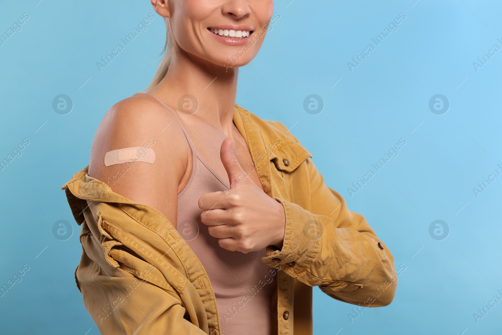 Photo of Smiling woman with adhesive bandage on arm after vaccination showing thumb up on light blue background, closeup