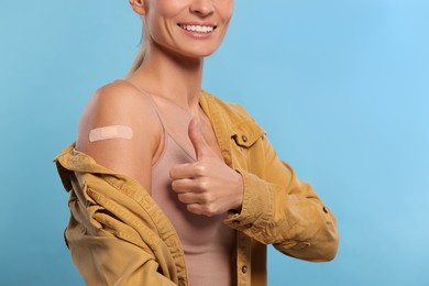 Smiling woman with adhesive bandage on arm after vaccination showing thumb up on light blue background, closeup