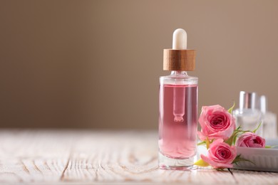 Bottle of essential rose oil and flowers on white wooden table against beige background, space for text