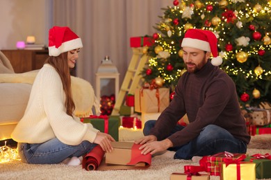 Happy couple in Santa hats decorating Christmas gift with wrapping paper at home