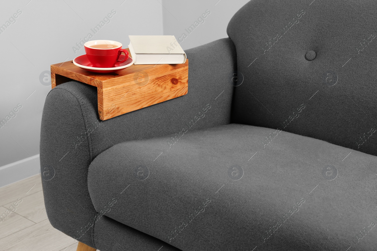 Photo of Cup of tea and book on sofa with wooden armrest table in room. Interior element
