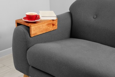 Cup of tea and book on sofa with wooden armrest table in room. Interior element