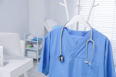 Photo of Blue medical uniform and stethoscope hanging on rack in clinic, closeup. Space for text