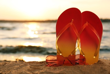 Stylish flip flops with sunglasses on sand near sea, space for text. Beach accessories