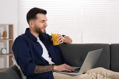 Photo of Handsome man with delicious smoothie using laptop on sofa at home