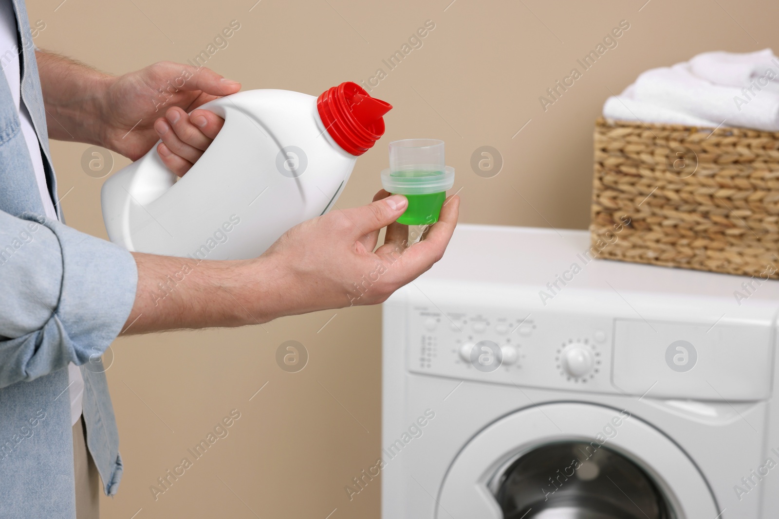 Photo of Man pouring fabric softener from bottle into cap near washing machine indoors, closeup