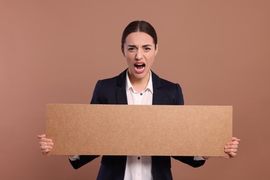 Photo of Angry young woman holding blank cardboard banner on brown background, space for text