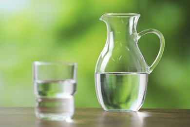 Photo of Jug and glass with clear water on wooden table against blurred green background, selective focus