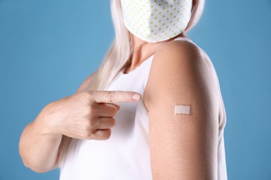 Mature woman in protective mask pointing at arm with bandage after vaccination on light blue background, closeup