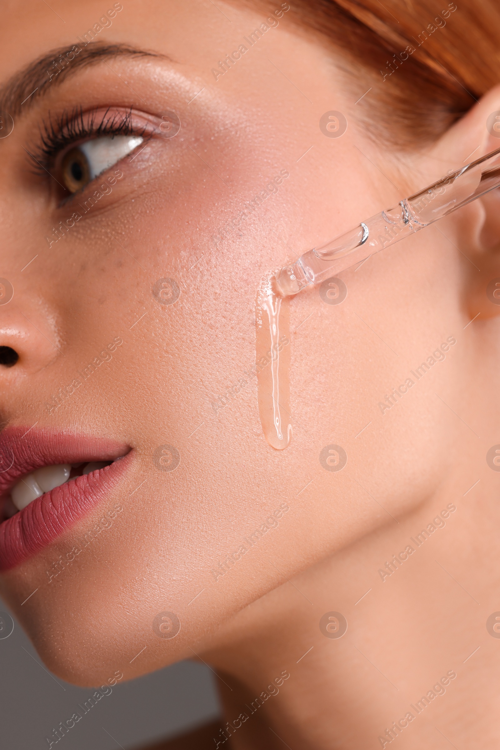 Photo of Beautiful young woman applying cosmetic serum onto her face on grey background, closeup