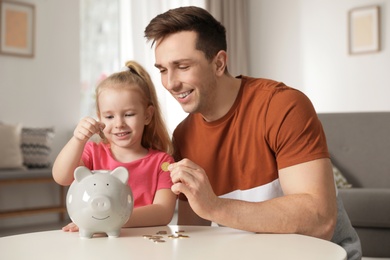 Photo of Father and daughter putting coins into piggy bank at table indoors. Saving money