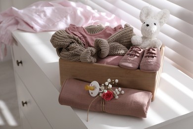 Wooden crate with children's clothes, shoes, toy and pacifier on chest of drawers in room