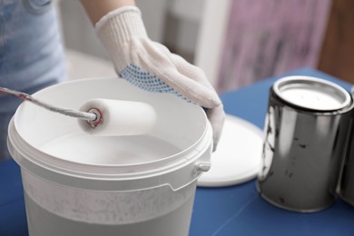 Woman dipping roller into bucket with white paint at blue wooden table indoors, closeup. Space for text