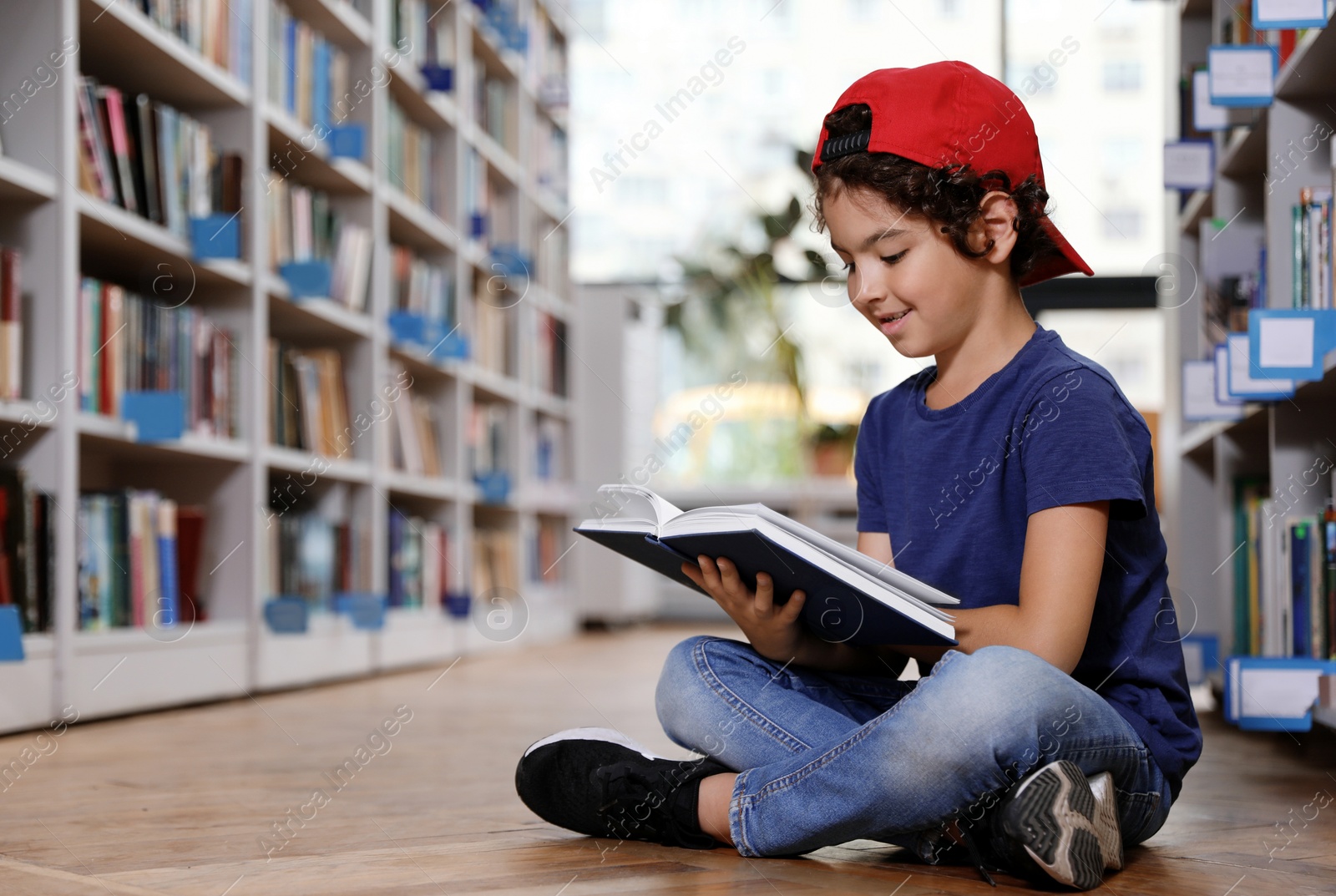 Photo of Cute little boy reading book on floor in library