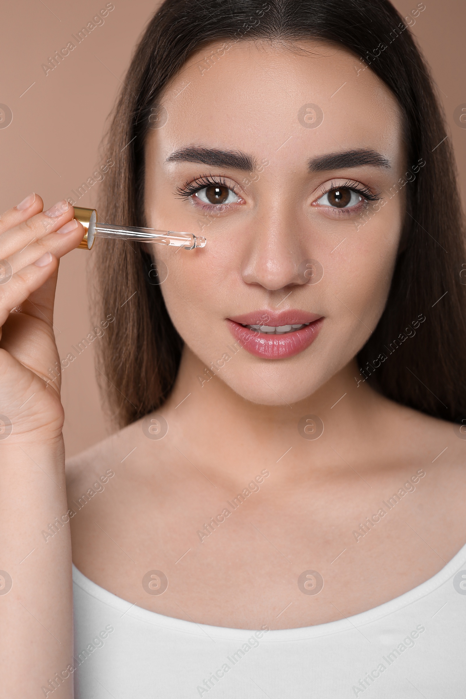 Photo of Woman applying essential oil onto face on brown background, closeup