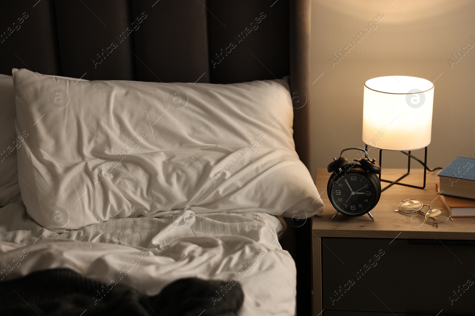 Photo of Nightlight, alarm clock, glasses and books on bedside table near bed indoors