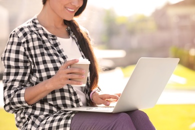 Woman with cup of coffee using laptop outdoors, closeup
