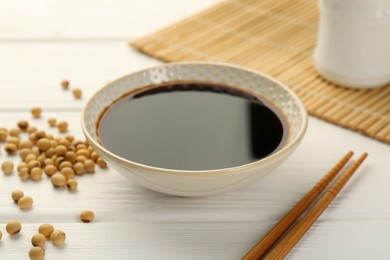 Photo of Tasty soy sauce in bowl, chopsticks and soybeans on white wooden table, closeup