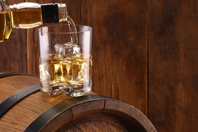 Pouring whiskey from bottle into glass on barrel against wooden background, closeup. Space for text