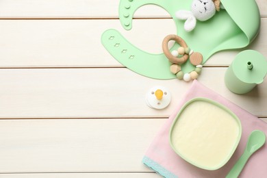 Photo of Silicone baby bib, toys and plastic dishware with healthy food on white wooden table, flat lay. Space for text