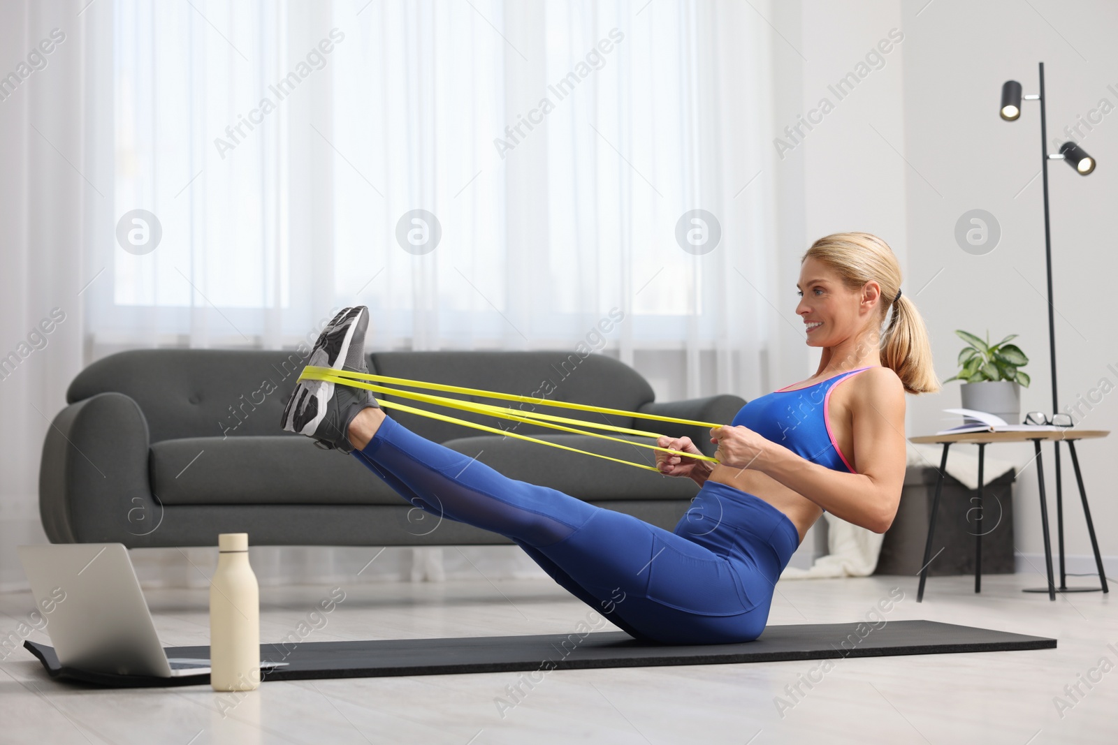 Photo of Fit woman doing exercise with fitness elastic band near laptop on mat at home