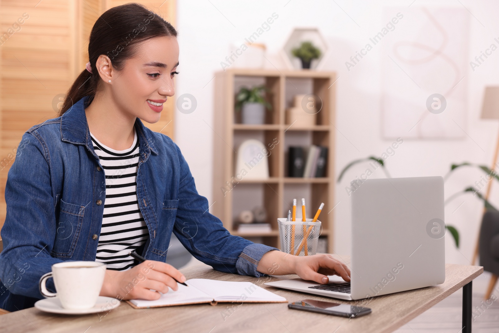 Photo of Young woman writing in notebook while working on laptop at wooden table indoors