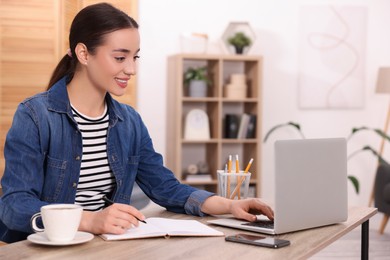 Young woman writing in notebook while working on laptop at wooden table indoors