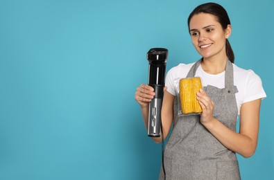 Photo of Beautiful young woman holding sous vide cooker and corn in vacuum pack on light blue background. Space for text