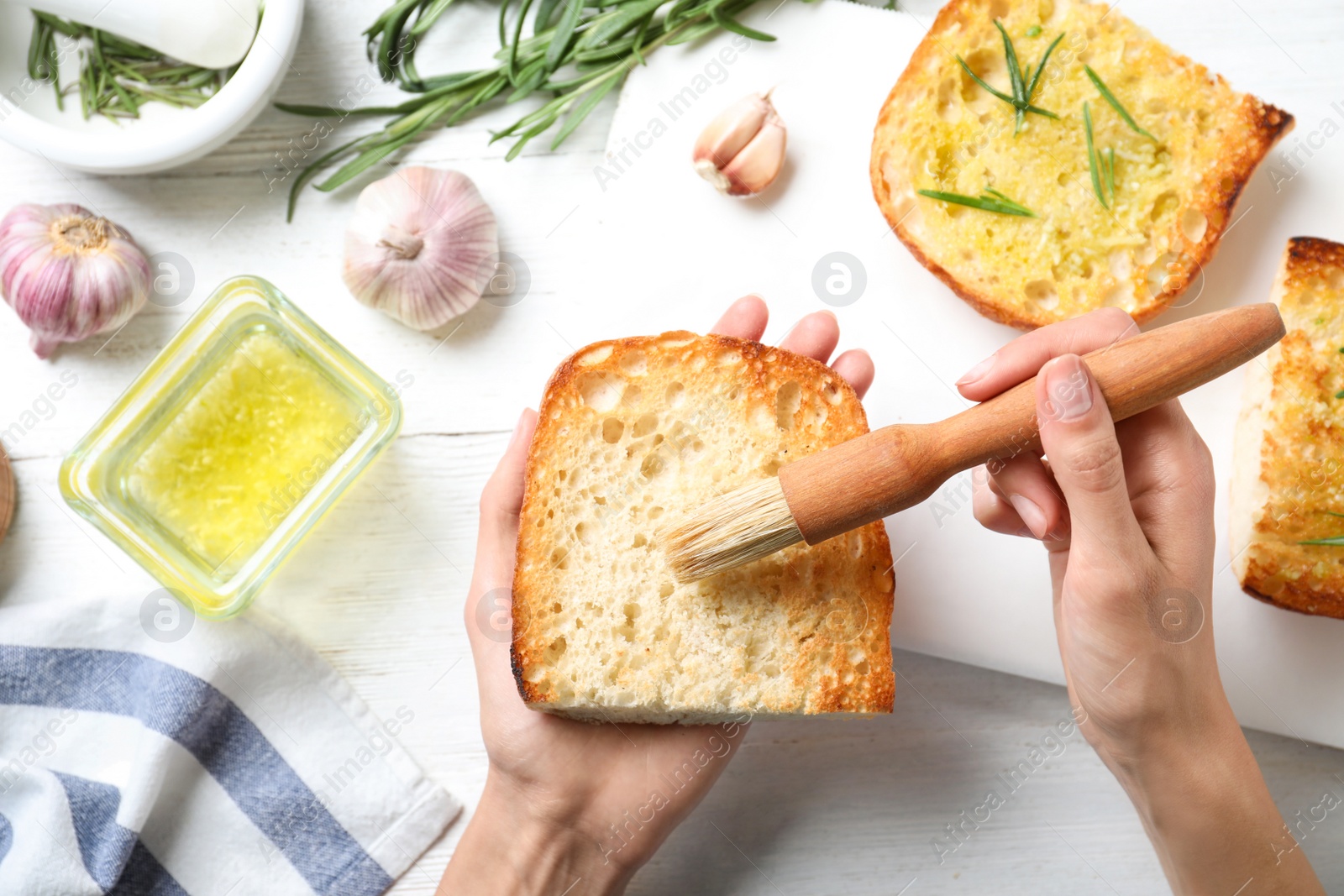 Photo of Woman brushing slice of toasted bread with garlic and herb over white wooden table, top view