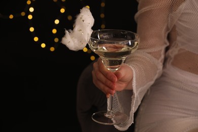 Woman holding glass of cocktail decorated with tasty cotton candy on black background with blurred lights, closeup. Space for text