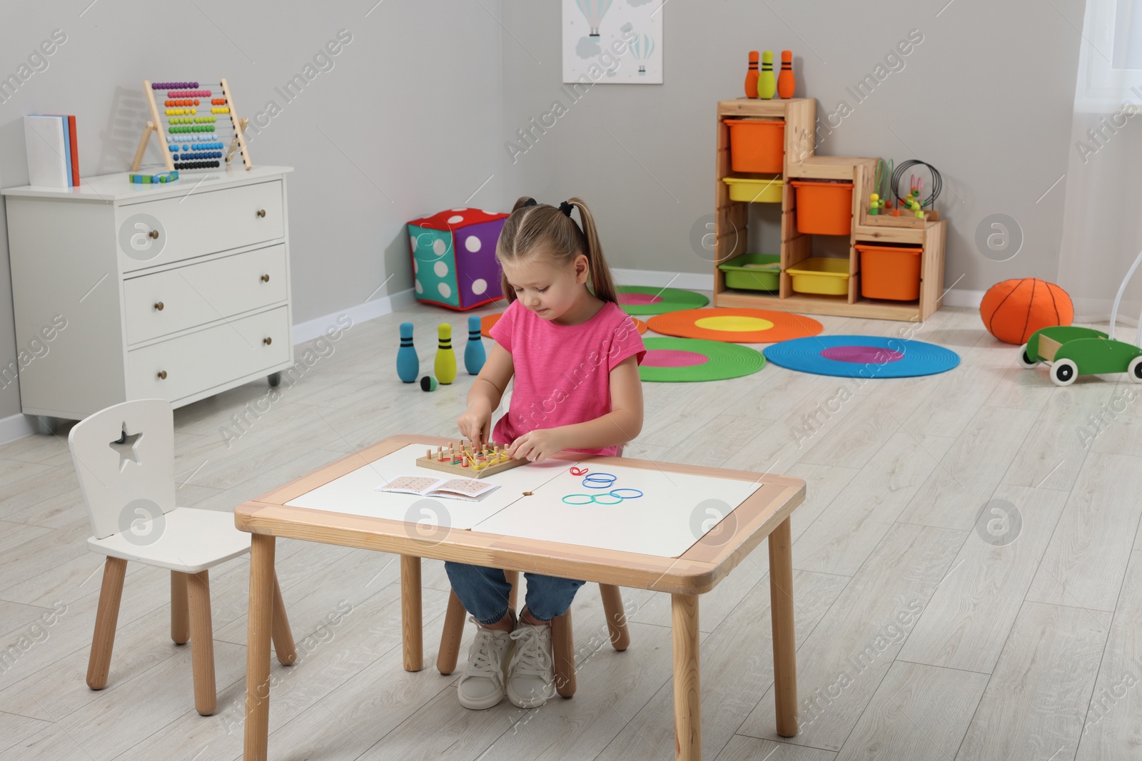 Photo of Motor skills development. Girl playing with geoboard and rubber bands at white table in kindergarten