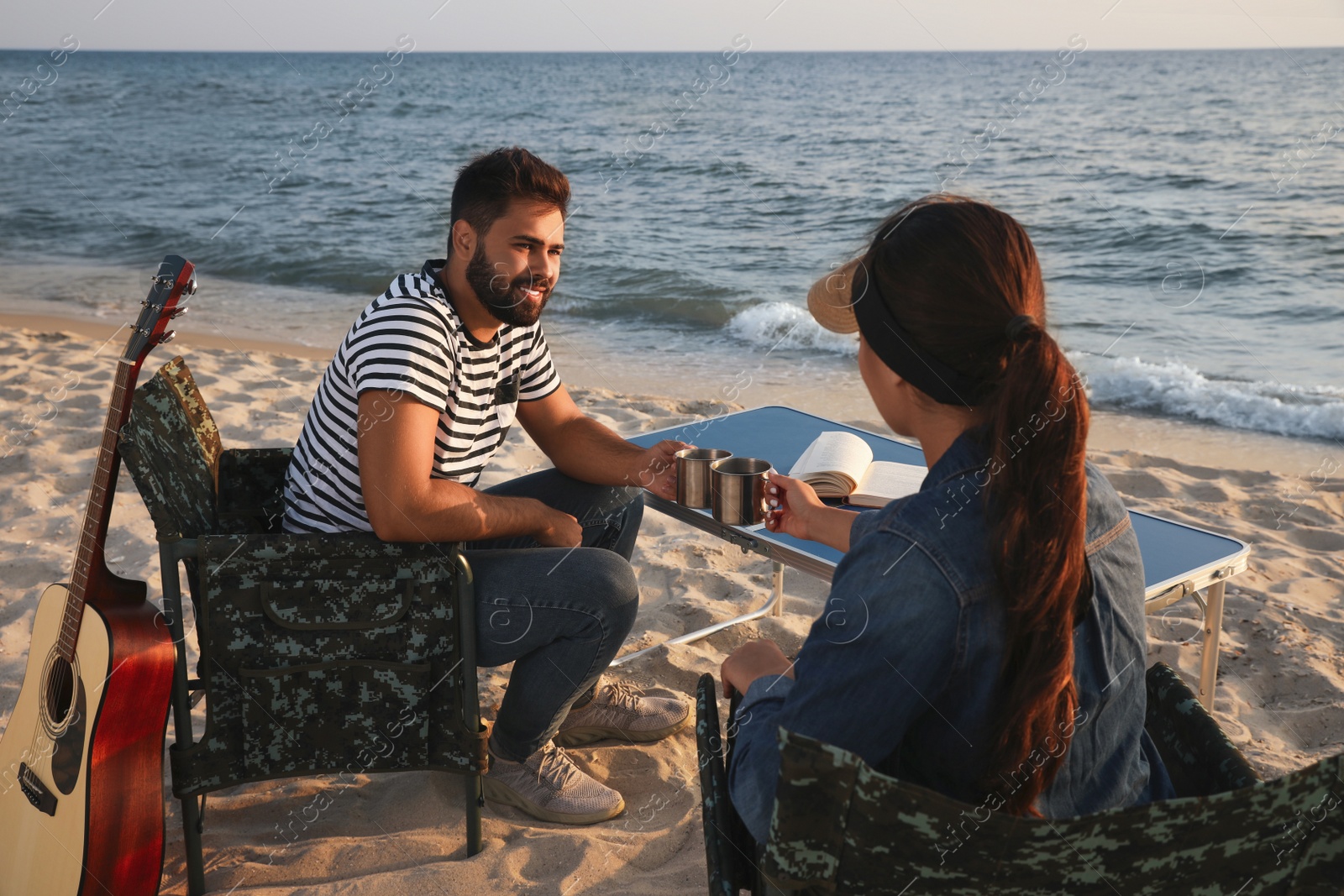 Photo of Couple sitting in camping chairs and clinking mugs on beach