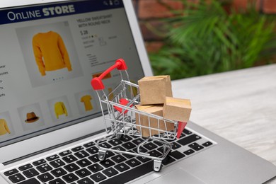 Photo of Internet store. Small cardboard boxes, shopping cart and laptop on light wooden table, closeup