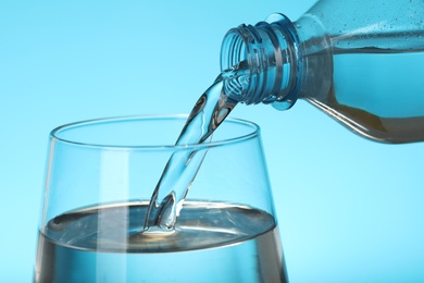 Pouring water from bottle into glass on blue background, closeup. Refreshing drink
