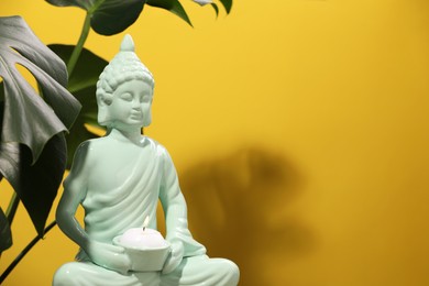 Photo of Buddhism religion. Decorative Buddha statue with burning candle and monstera against yellow wall, space for text
