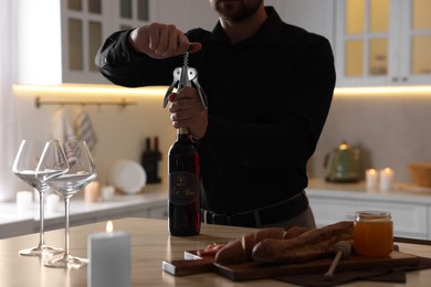 Photo of Romantic dinner. Man opening wine bottle with corkscrew at table in kitchen, closeup