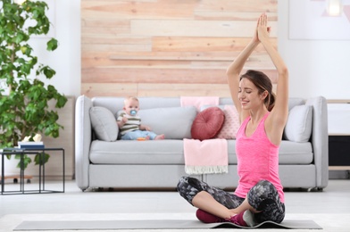 Young sportive woman doing exercise while her son sitting on sofa at home, space for text. Fitness training