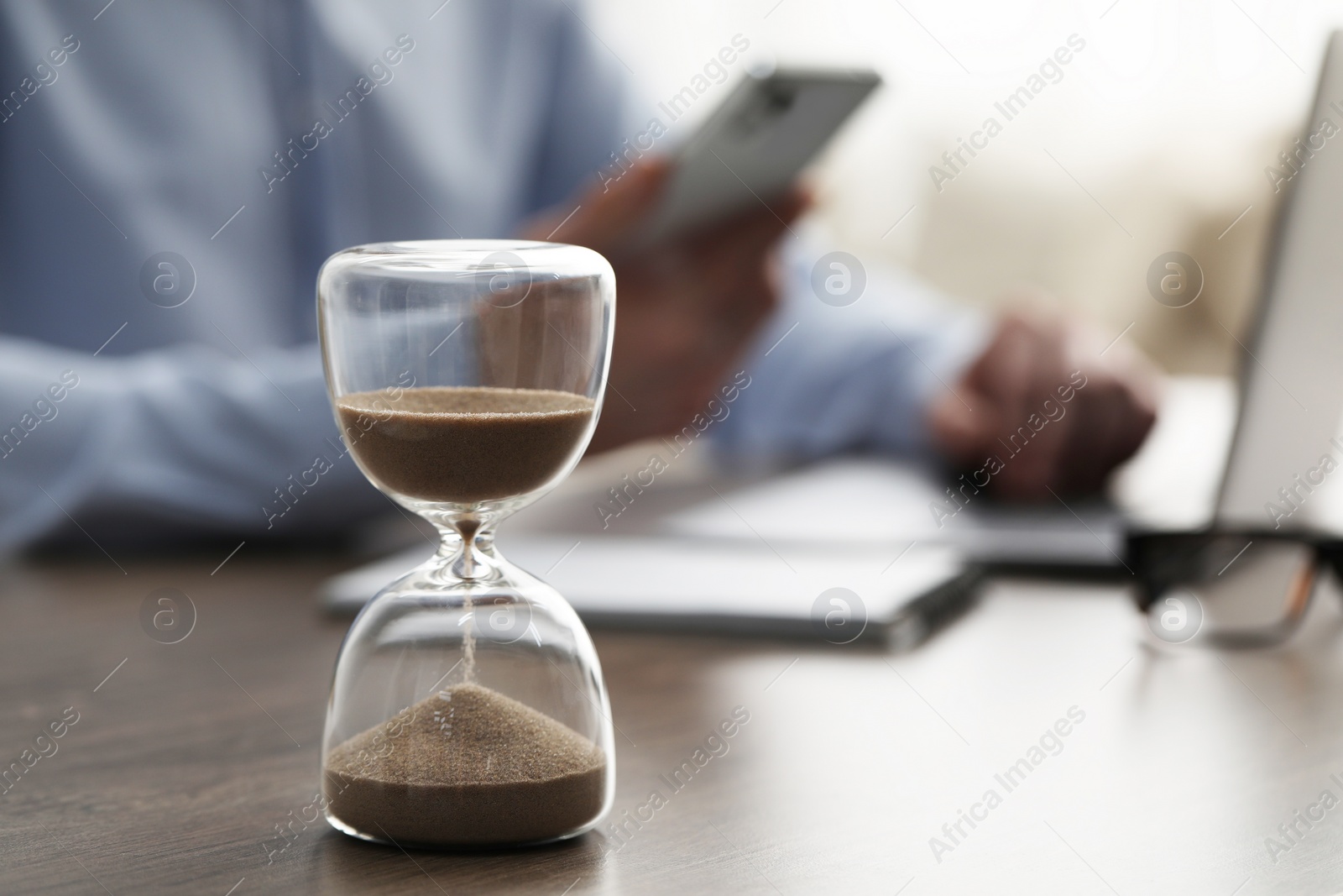 Photo of Hourglass with flowing sand on desk. Man using smartphone while working on laptop indoors, selective focus