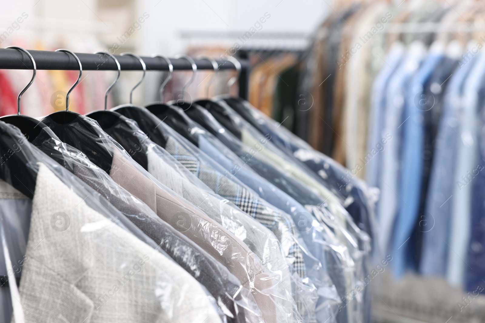 Photo of Dry-cleaning service. Many different clothes in plastic bags hanging on rack indoors, closeup