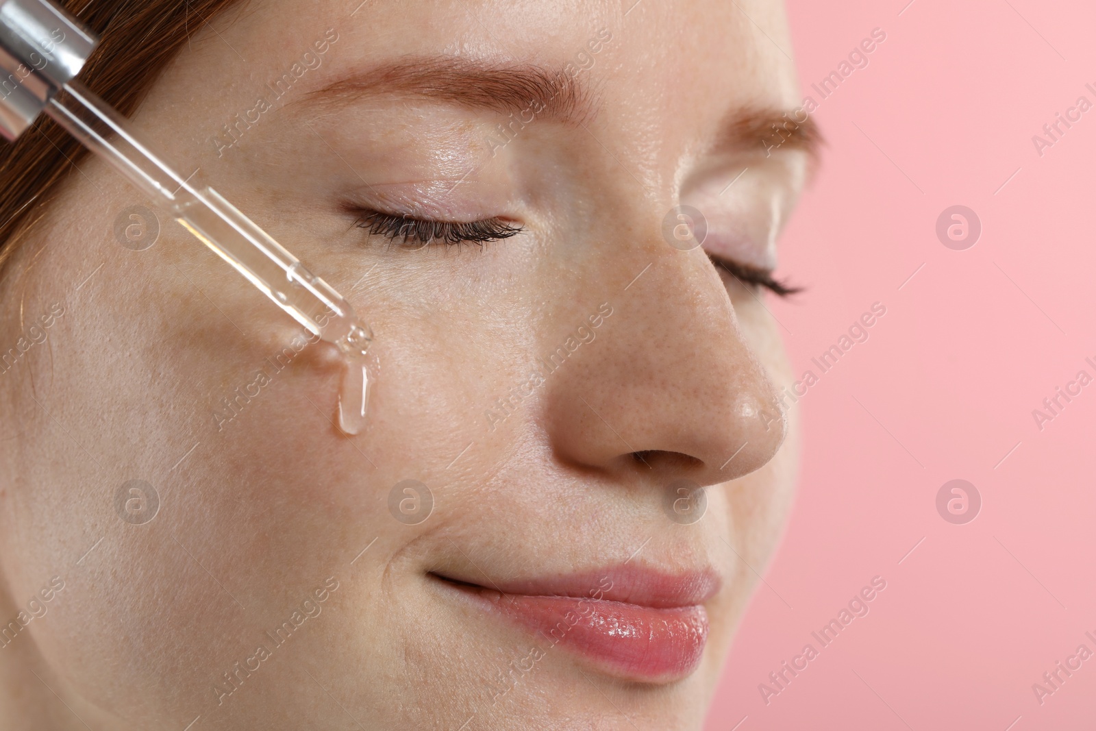 Photo of Beautiful woman with freckles applying cosmetic serum onto her face on pink background, closeup