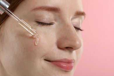 Photo of Beautiful woman with freckles applying cosmetic serum onto her face on pink background, closeup