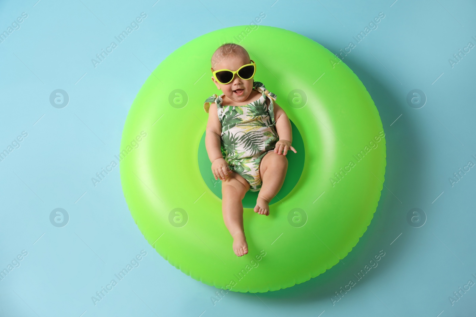Photo of Cute little baby in sunglasses with inflatable ring on light blue background, top view